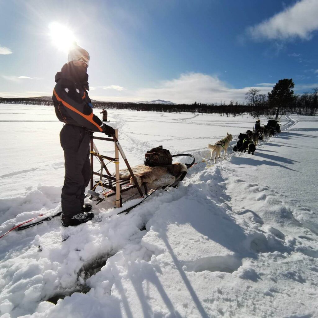 Una persone in tuta da sci poggia le mani su una slitta trainata da husky in mezzo alla neve