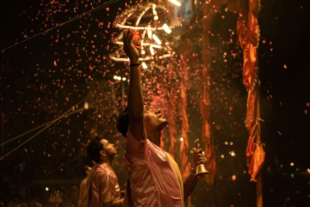 Sacerdote indù durante una cerimonia serale sul Gange, solleva una mano tra petali e fumo rituale.